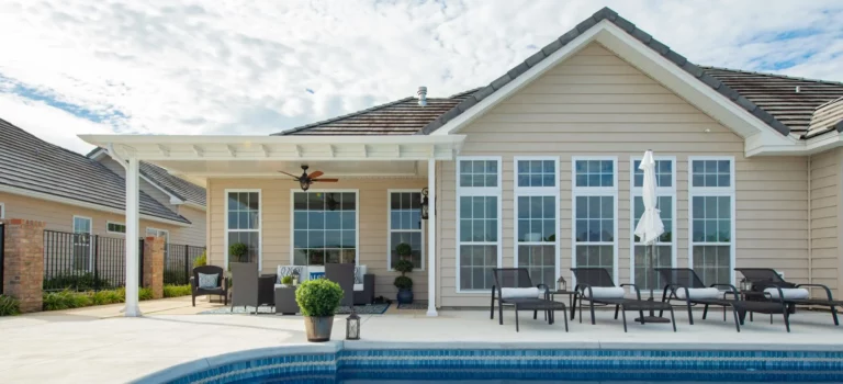 A modern house exterior with a covered patio, featuring wicker seating and potted plants, alongside a pool with a blue tiled edge. The house has large windows with multiple panes and a sloped roof under a partially cloudy sky.