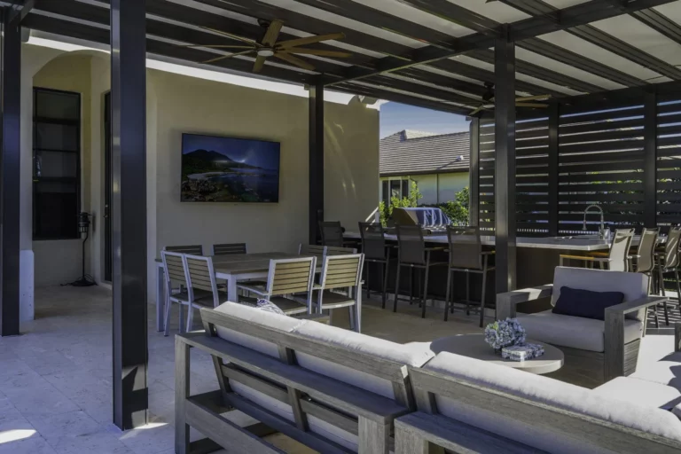 Cooking in Your Outdoor Living Space 7 A modern outdoor patio area featuring a seating arrangement with gray sofas, a coffee table, and dining tables with chairs. There is a large flat-screen TV mounted on a wall, a ceiling fan above, and a bar-style seating area with high chairs in the background. The space is partially shaded and has a clean, contemporary design.