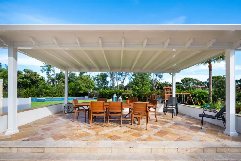 A spacious outdoor dining area under a white pergola, featuring a wooden table with several chairs, situated on a patio with stone flooring. In the background, there are trees, a grassy area, and a playground with swings, along with a blue swimming pool visible beyond a white fence.