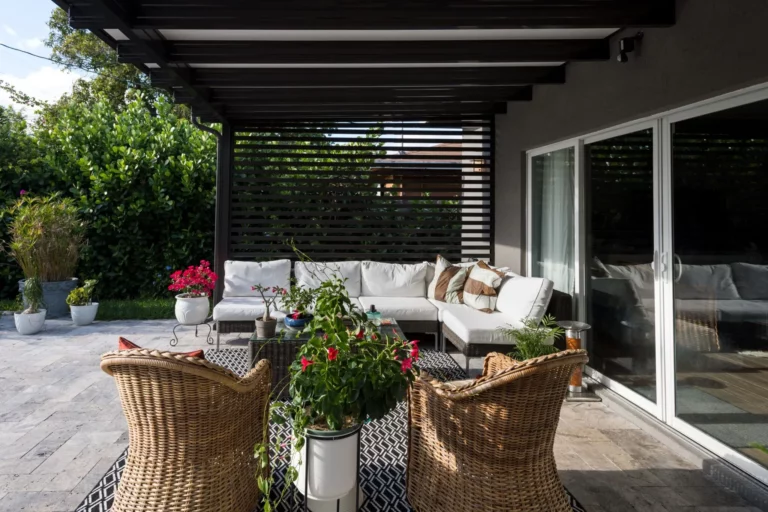 Furnishing your new patio 9 A cozy outdoor seating area featuring a white sectional sofa with decorative pillows, wicker chairs, and various potted plants. The space is partially shaded by a slatted overhead structure, with a stone floor and green foliage in the background.