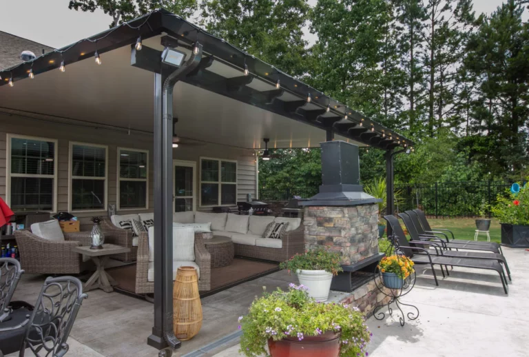How to Choose the Right Patio Builder in Richmond for Your Project 11 A covered outdoor patio with a stone fireplace, comfortable seating arranged around a coffee table, and potted plants, surrounded by greenery and a fence. Decorative string lights hang from the ceiling.
