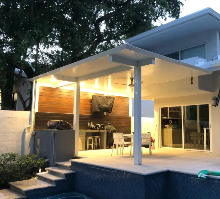 Insulated Patio Roofing: Advantages and Drawbacks 3 A modern outdoor patio with a covered seating area featuring a dining table and chairs, a grilling station with a black cover, and illuminated wood paneling. There are steps leading down to a pool area, and trees provide natural shading in the evening light.