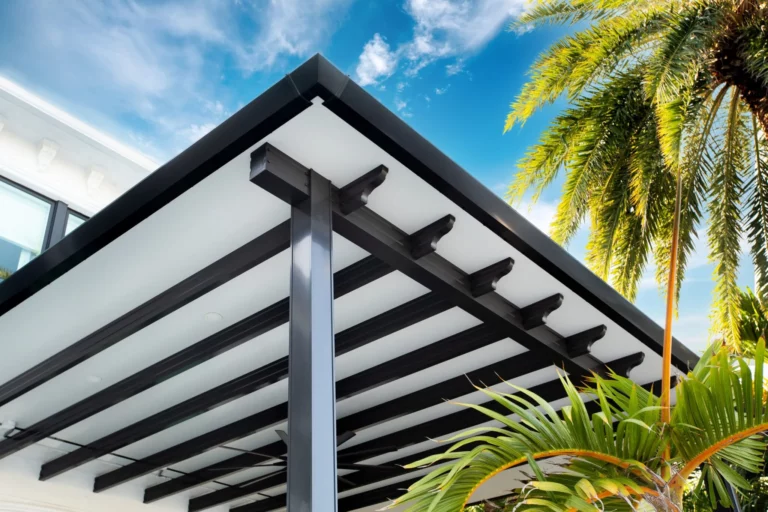 A close-up view of a modern outdoor patio roof with black beams and a white ceiling, alongside lush green palm leaves under a bright blue sky with scattered clouds.