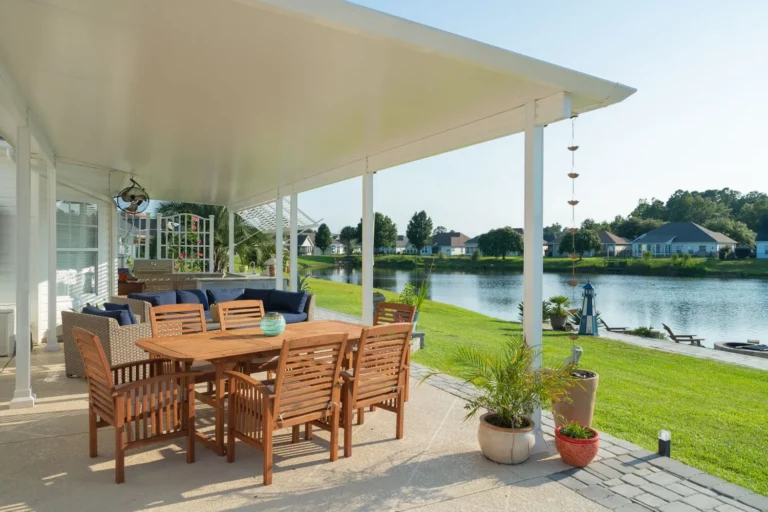 A covered outdoor patio with a wooden dining table and chairs, a blue sofa, and decorative plants, overlooking a calm lake and green lawn, with houses visible in the background.
