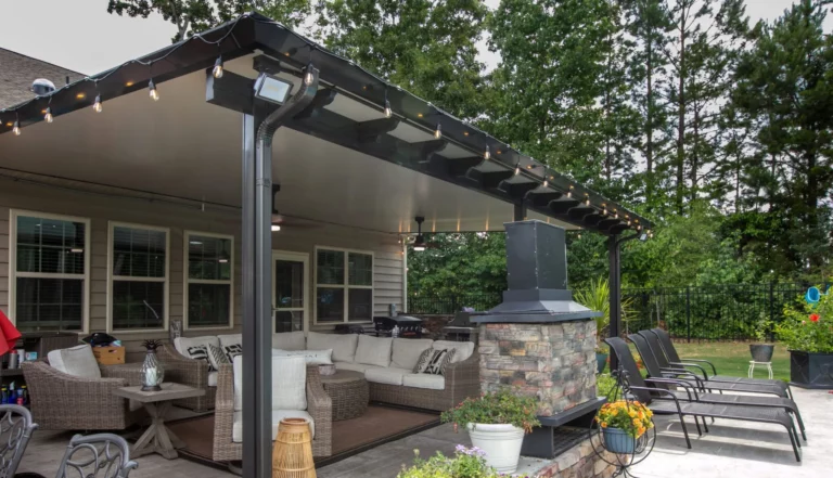 A covered outdoor patio area featuring comfortable seating, a stone fireplace, and decorative plants, with string lights hanging from the roof and lush greenery in the background.