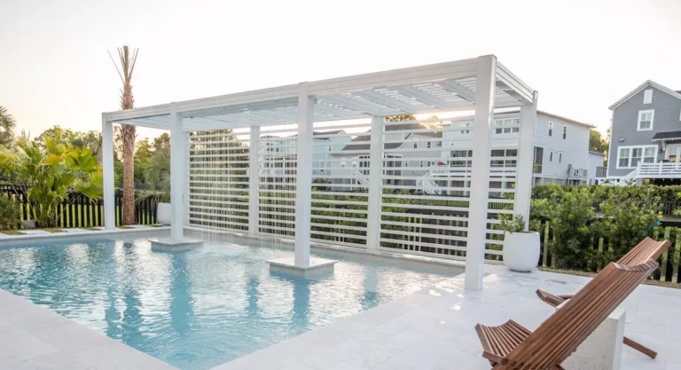 A serene backyard with a swimming pool reflecting the sunset, featuring a modern white pergola with vertical slats, surrounded by greenery and a wooden lounge chair.