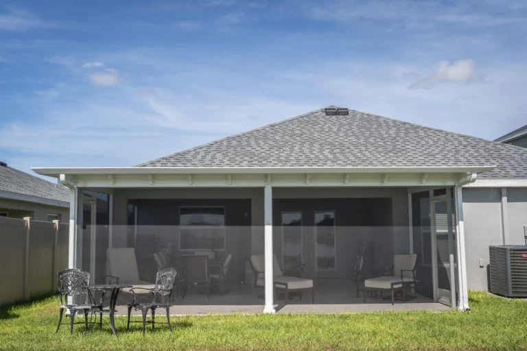 A view of a modern backyard patio with a gray shingled roof, featuring a screened area with a table and chairs, surrounded by green grass under a bright blue sky.